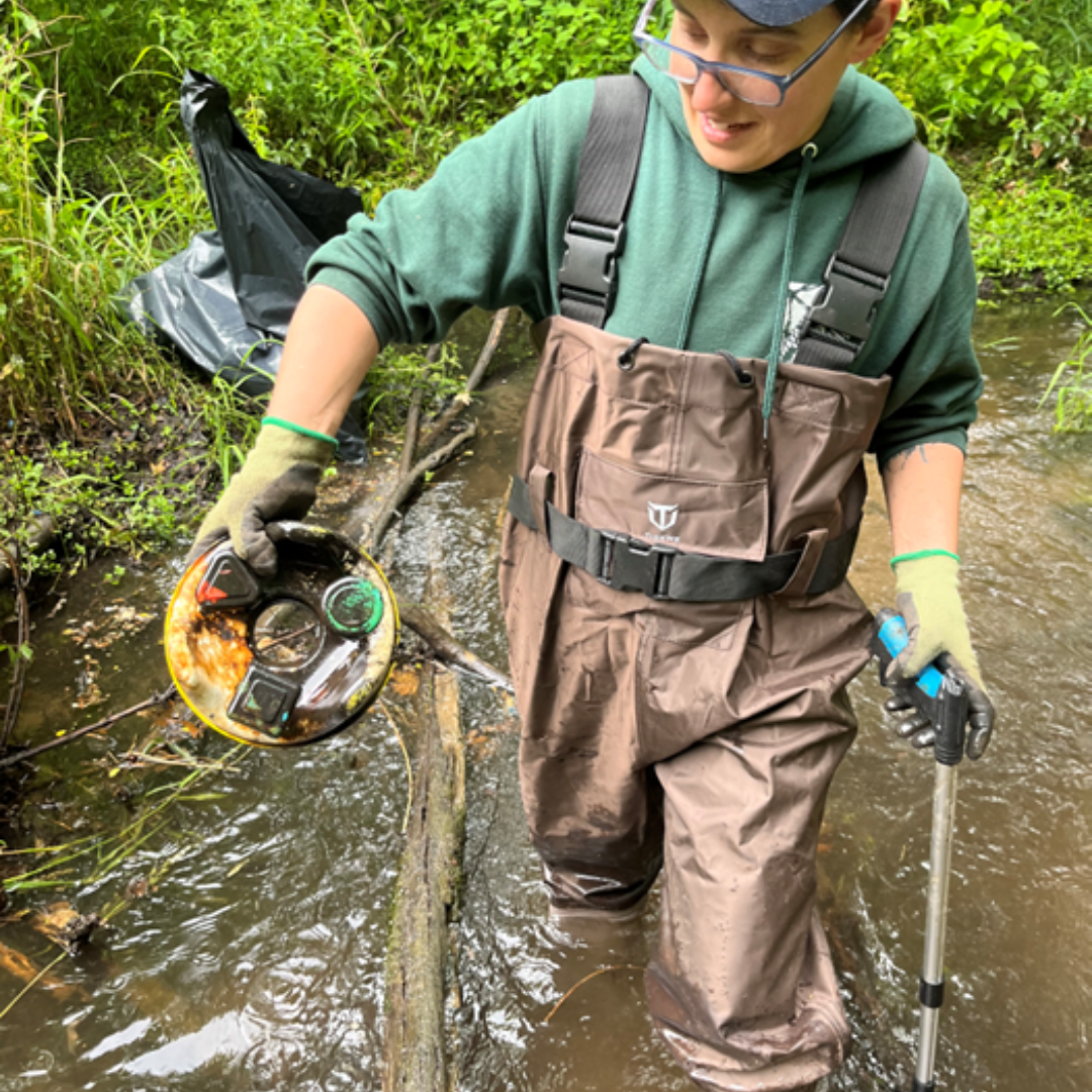 Holding trash, wearing waders, in creek
